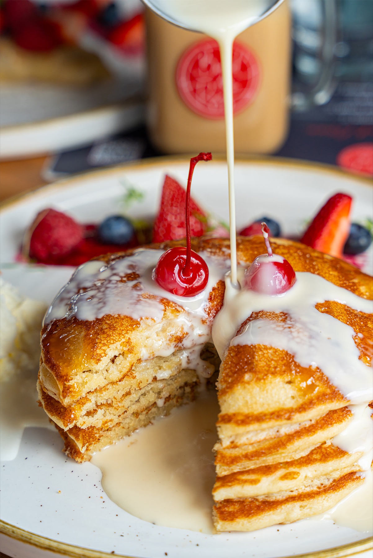 Cream poured over a cut stack of pancakes with cherries and berries
